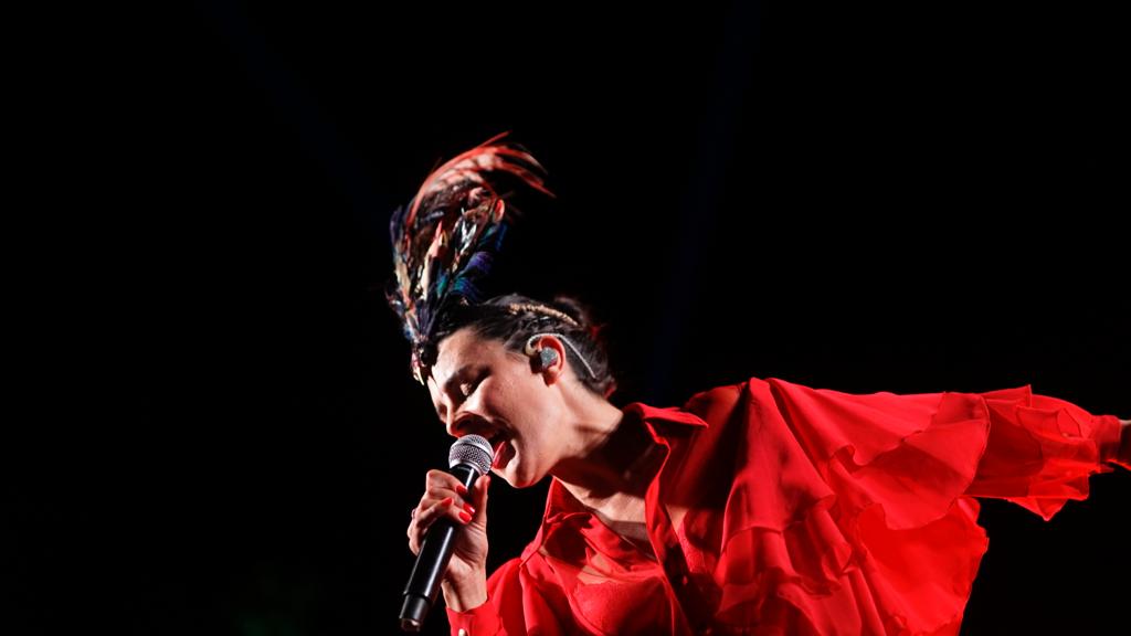 Fuel Fandango canta con una camisa roja, pelo recogido y decorado sobre un fondo negro